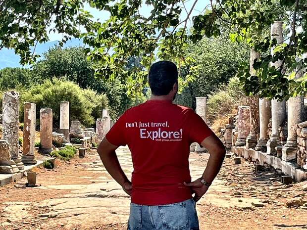 Person stands with hands on hips, wearing a red shirt reading "Don't just travel. Explore!" amidst ancient ruins with columns, under leafy trees and a clear sky.